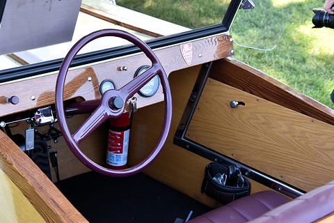 Purple steering wheel, a small round speedometer on a wooden dash, a red fire extinguisher and a varnished wooden door panel. Opens a larger view