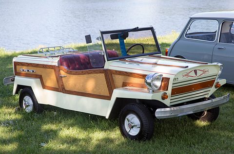 Cream open two-seater with woodgrain side panels, red seats and a small windscreen, parked on grass by water, a pale blue saloon behind.