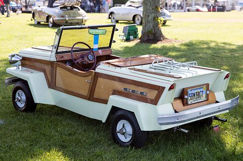 Rear quarter of a cream open two-seater with woodgrain panels, a chrome luggage rack on the tail and a chrome bumper, on a lawn. Opens a larger view