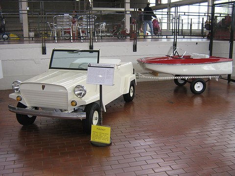 White open two-seater with a small windscreen and an information sign, beside a white and red dinghy on a trailer, on a brick floor.