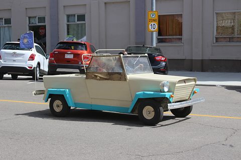 Small cream open roadster with turquoise wings and wheel arches, its folded top down, on a street in front of a low commercial building. Opens a larger view