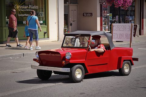 Red open two-seater with a black top and white wheels driven along a street, people walking on the pavement in front of shops. Opens a larger view