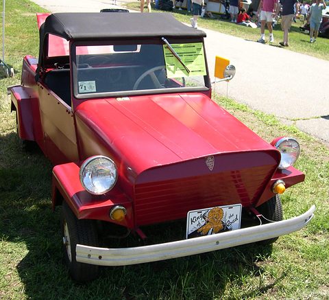 Low front view of a red open two-seater with a black top, round headlamps, a slatted grille and a chrome bumper, on a lawn. Opens a larger view