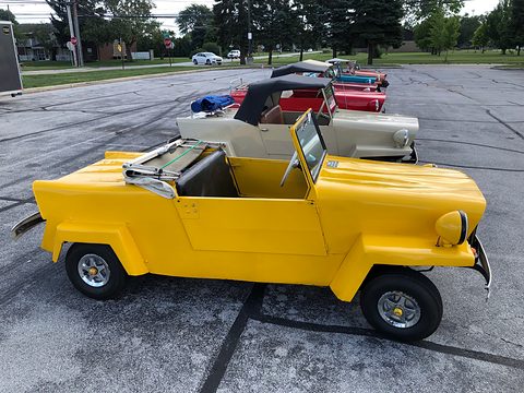 Bright yellow open two-seater with a folded windscreen and doors, in profile on a car park, a line of similar cars behind. Opens a larger view