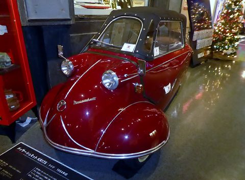 Red tandem microcar with black soft top, a Japanese label board in front. Opens a larger view