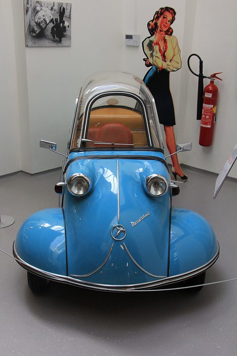Bright blue bubble car head-on in a white room, a pin-up cut-out and fire extinguisher behind. Opens a larger view