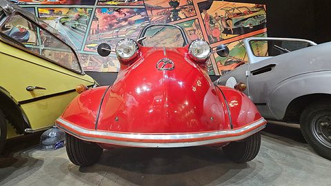 Red bubble car head-on with two headlamps on its nose, a comic-strip mural behind. Opens a larger view