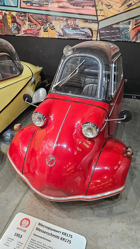 Red tandem microcar seen from above at the front quarter, a museum label beside it. Opens a larger view