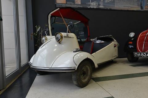 A white three-wheeled cabin scooter with two headlamps close together on its nose and a red-lined canopy swung up. Opens a larger view