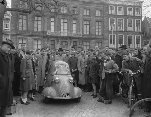 Men and boys in coats press round a small tandem-seat bubble car on a cobbled street.