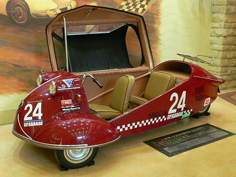 Red open tandem microcar with number 24 decals and a chequered stripe in a museum. Opens a larger view