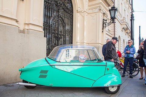 Turquoise bubble car side-on beside an ornate doorway, a few people talking behind it. Opens a larger view