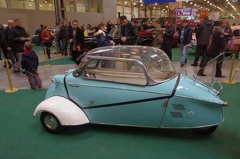 Turquoise bubble car with white wings on green carpet, show visitors behind. Opens a larger view