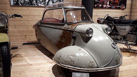 Grey bubble car with rust patches on a wooden floor among motorcycles. Opens a larger view