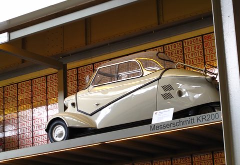 Cream bubble car on a shelf against a wall of old number plates, labelled Messerschmitt KR200. Opens a larger view