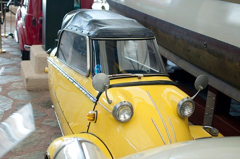 Close view of a yellow three-wheeler’s nose with two headlamps and mirrors under a black soft top. Opens a larger view