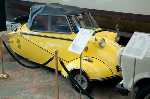 Yellow three-wheeler with a black soft top behind a rope barrier, information sheets on stands. Opens a larger view