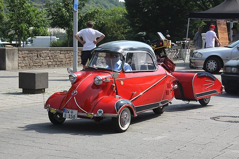A red 1955 KR200 with a black canopy roof, towing a small matching red trailer across a car park. Opens a larger view