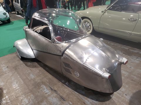 Bare aluminium-coloured bubble car with visible panel joints on a grey stand. Opens a larger view