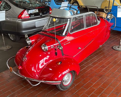 Red three-wheeler with folded soft top and chrome bumper bars on a brick floor. Opens a larger view