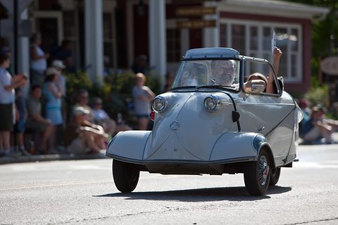 Grey bubble car with canopy open, driver and passenger aboard, spectators along the street. Opens a larger view
