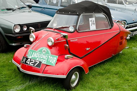 Red three-wheeler with black soft top and a green rally plate on its nose among parked cars. Opens a larger view
