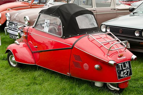 Red three-wheeler with black soft top and chrome rack, number 74 in the window, on grass. Opens a larger view