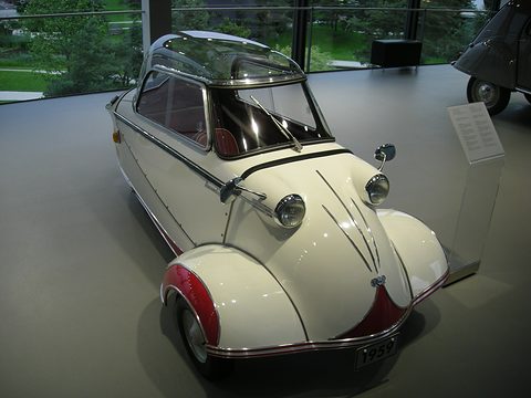 White bubble car with red wheel arches on a grey floor beside an information stand. Opens a larger view