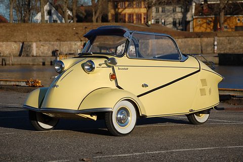 Cream three-wheeler with dark soft top in low evening light on a waterside road. Opens a larger view