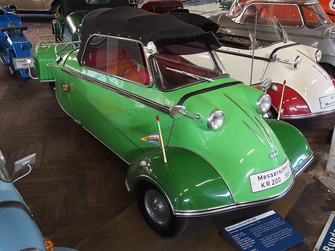 Green three-wheeler with black soft top among other Messerschmitts on a parquet floor. Opens a larger view