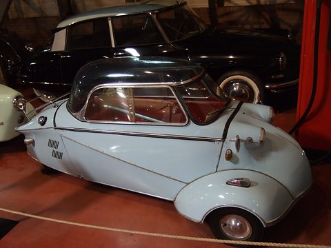 Light blue three-wheeler with a dark hard top behind a rope, a black saloon behind. Opens a larger view