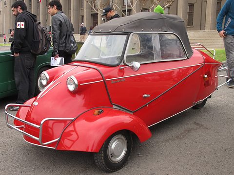 Red three-wheeler with a black soft top and chrome bumper bar, people standing around it. Opens a larger view