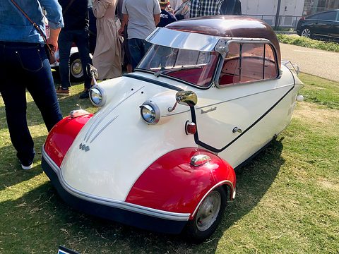 Red and white bubble car with a maroon-tinted canopy on grass, visitors’ legs behind. Opens a larger view