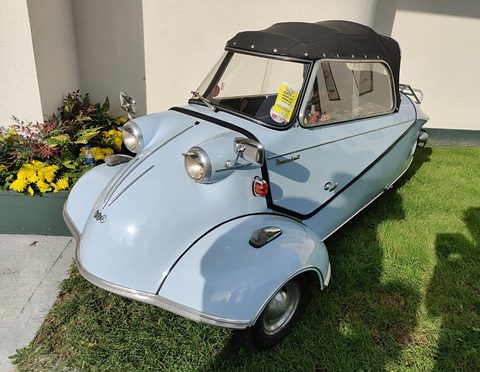 Pale blue three-wheeler with black soft top on a lawn beside a yellow flower bed. Opens a larger view