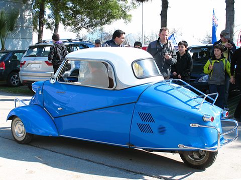 Blue three-wheeler with a white hard top and chrome rack, seen from the rear quarter, visitors behind. Opens a larger view