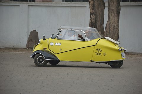 Yellow tandem bubble car with black wings driving past a white wall. Opens a larger view