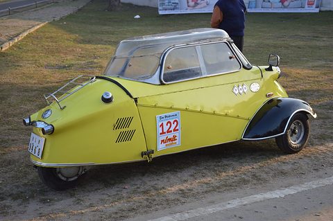 Yellow three-wheeler with black wings and rally plates, seen from the rear quarter on a dusty ground. Opens a larger view