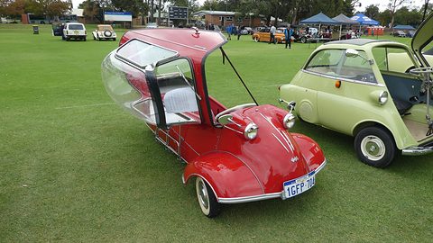 Red three-wheeler with its canopy swung up beside a pale green Isetta on a playing field. Opens a larger view