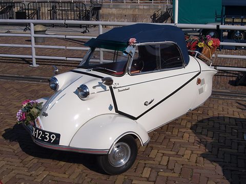 White three-wheeler with navy soft top, flowers on the nose, parked on a brick quay. Opens a larger view