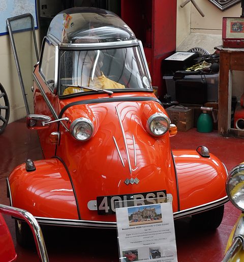 Red bubble car head-on in a garage, an information card propped in front. Opens a larger view