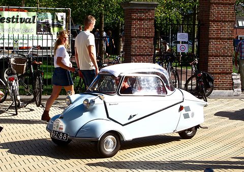 Light blue and white bubble car with its driver moving off past a gate, two pedestrians behind. Opens a larger view