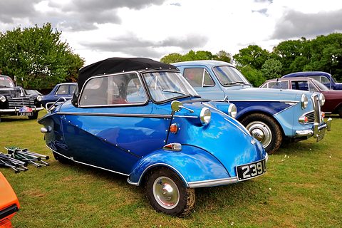 Metallic blue three-wheeler with black soft top on grass among classic saloons. Opens a larger view