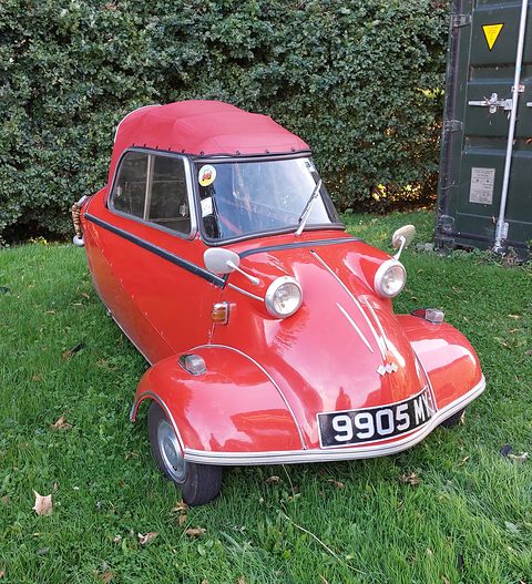 Red three-wheeler with red soft top on grass in front of a hedge. Opens a larger view