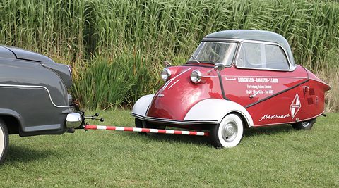 Maroon and white three-wheeler lettered Abholdienst, linked by a striped tow bar to a grey car.