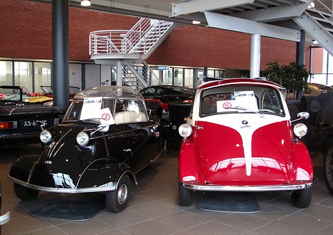 Black bubble car next to a red and white Isetta in a showroom. Opens a larger view