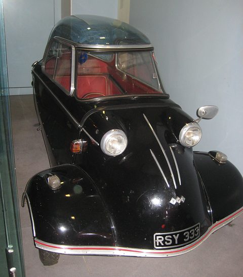 Black bubble car with red interior seen head-on inside a glass case. Opens a larger view