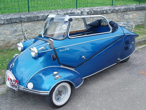 Metallic blue three-wheeler with canopy raised and a chrome rack beside a stone wall. Opens a larger view