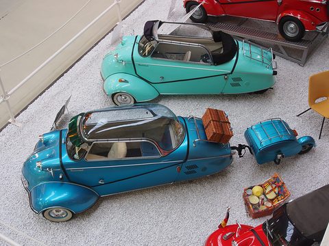 Blue bubble car with a brown case on its rack and a small blue trailer, seen from above on gravel. Opens a larger view