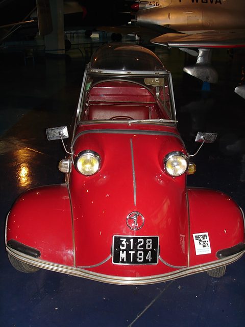 Red bubble car with registration 3128 MT 94 in a dim hangar, aircraft above. Opens a larger view