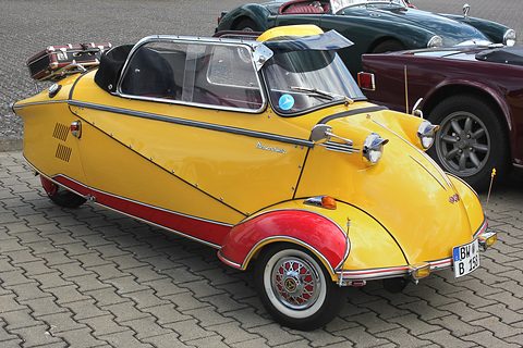 Yellow and red three-wheeler with folded soft top, whitewall tyres, on paving. Opens a larger view